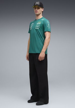 Giovane uomo con indosso una maglietta verde del team Aston Martin Aramco Formula Uno con loghi degli sponsor, pantaloni neri a gamba larga, cappellino e scarpe nere, in piedi.