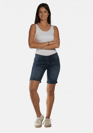 Shorts en denim bleu foncé avec une fermeture à cinq boutons, ourlet légèrement retroussé, associés à un haut sans manches blanc et des chaussures de couleur claire.