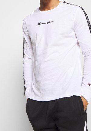 Persona que lleva una camiseta blanca de manga larga de la marca Champion con el logo en el pecho y franjas negras con el logo en las mangas, combinada con pantalones negros.