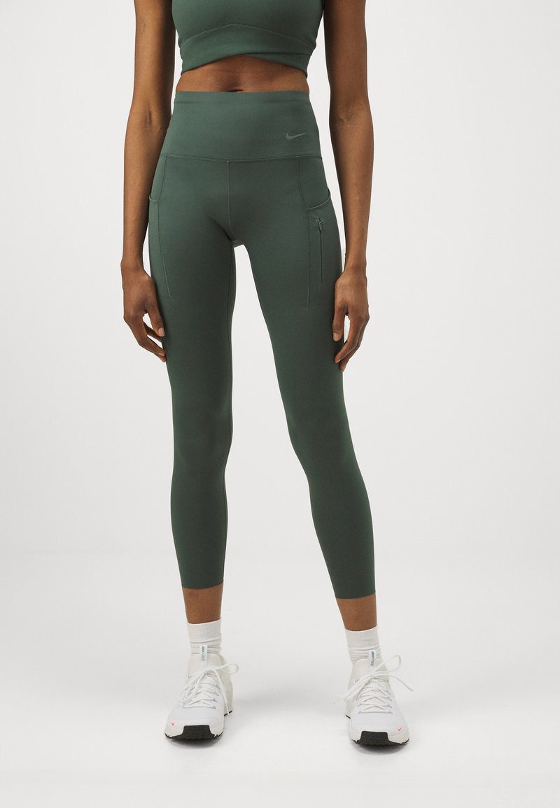 Grønne leggings med høy livvidde, sidelommer med glidelås og glatt, elastisk stoff. Brukt med hvite treningsko og ankelsokker.