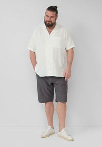 Man met een baard en teruggebonden haar, gekleed in een witte short-sleeve button-up shirt, donkere gestreepte korte broek en witte sneakers, staand tegen een effen muur.