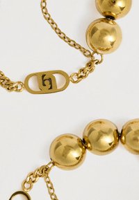 Zlatý náramek na řetízku se velkými, lesklými kulatými korálky a bezpečnou sponou se značkou. Design spojuje hladké a texturované prvky.
