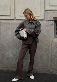 Vrouw in een donkerbruine leren jas en leren broek, met een kleine witte handtas, staande tegen een gestructureerde stenen muur.