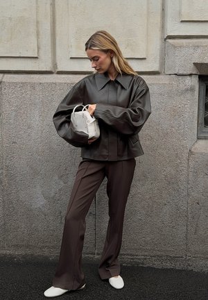 Vrouw in een donkerbruine leren jas en leren broek, met een kleine witte handtas, staande tegen een gestructureerde stenen muur.