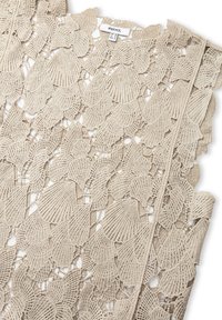 Tissu en dentelle beige avec un motif de feuilles florales présentant des détails intricats et des bords festonnés ; étiquette visible au niveau du cou.