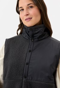 Schwarz isolierte Weste mit Fleece- und Stoffpaneelen, hohem Kragen, Druckknöpfen und Markenlogo auf der linken unteren Seite.