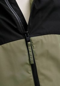 Oliivinvihreä ja musta vetoketjullinen takki, jossa on teksturoitu pinta. Takissa on tukeva vetoketju ja värilohkottu muotoilu rintakehän alueella.