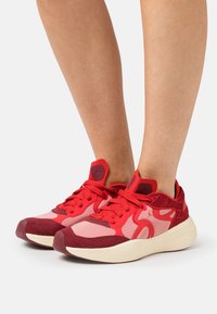 Pieds portant des baskets de sport rouges et roses avec des semelles blanches, se tenant sur un fond clair.