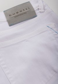 White fabric pants with a gray leather label reading "bugatti HANDCRAFTED," featuring blue stitching details and a simple pocket design.