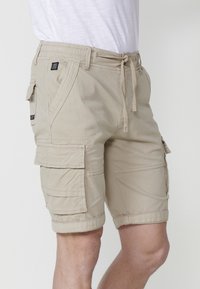 Pantaloni scurți cargo bej cu talie elastică și șnur, având multiple buzunare laterale și detalii cusute. Textură a țesăturii moale.