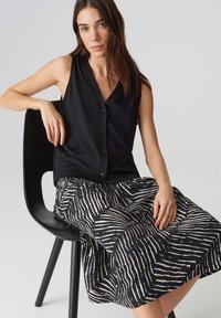 Mustat hihattomat paita napilla yhdistettynä musta-valkoiseen kuviolliseen hameeseen, jossa on abstrakteja raitoja, istuen mustassa tuolissa.