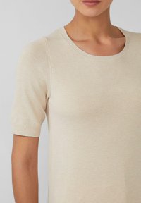 Beige Kurzarmpullover mit rundem Ausschnitt. Aus weichem Strickmaterial gefertigt, mit glatter Textur und dezenten Rippenelementen an den Ärmeln und dem Ausschnitt.