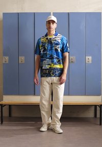 Korte mouwen, blauwe gepatterneerde polo met gele accenten, gecombineerd met crèmekleurige broeken en witte sneakers. Kluisjes op de achtergrond.