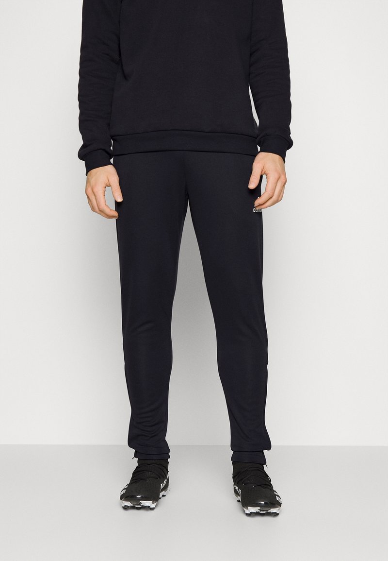Homme portant une chemise noire à manches longues, un pantalon noir ajusté et des chaussures de sport noires à crampons, debout devant un fond uni clair.