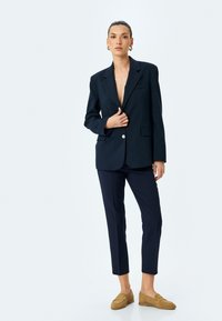 Blazer marine à bouton simple, avec des revers crantés et des poches avant, associé à un pantalon marine ajusté et des mocassins beiges. Texture lisse.