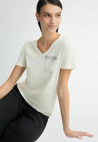Cremefarvet V-hals t-shirt lavet af blødt stof, med en lomme prydet med et rhinestones mønster. Sammen med sorte bukser udsmykket med glitrende detaljer.