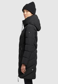 Zwarte pufferjas met een chevronpatroon, capuchon, zijzakken met rits en een logopatch. Een ribgebreide zwarte beanie maakt de outfit af.