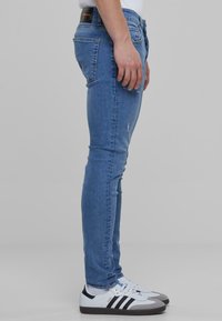 Kék vászonnadrág, slim fit szabással és megkopott részletekkel, fehér sportcipővel viselve, fekete kiemelésekkel.
