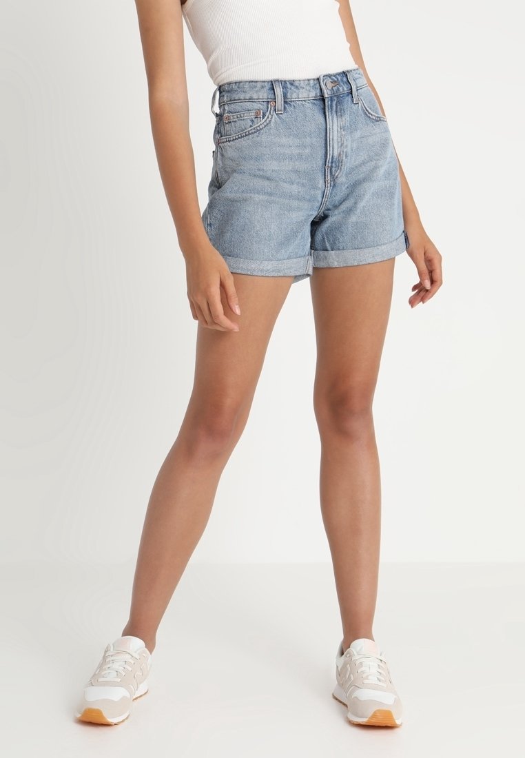 Shorts en denim bleu clair à taille haute, ourlets retroussés, et design à cinq poches. Associés à des baskets blanches, debout sur un fond uni.