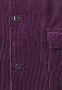 Kordtöjsfabric i djup lila med vertikala ribbor, två mörklila knappar och en ficklock på höger sida av plagget.