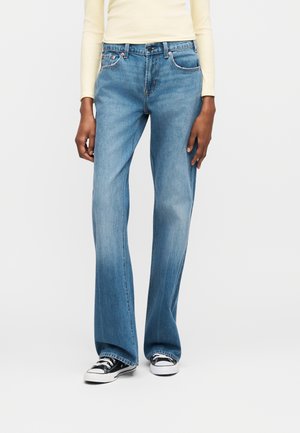 Άτομο με φαρδιά τζιν παντελόνια ανοιχτού μπλε χρώματος, μαύρα και λευκά αθλητικά παπούτσια, και μακρυμάνικη μπλούζα ανοιχτού κίτρινου χρώματος, στέκεται μπροστά σε μια απλή φόντο.