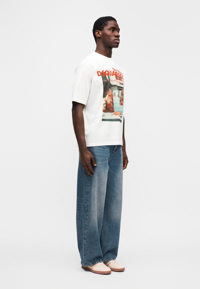 Λευκό γραφικό μπλο υζάκι με φωτογραφικό τύπωμα. Συνδυάζεται με φαρδιά τζιν παντελόνια μπλε ξεθωριασμένα και παπούτσια σε κρεμ χρώμα. Το μοντέλο στέκεται πλευρικά.