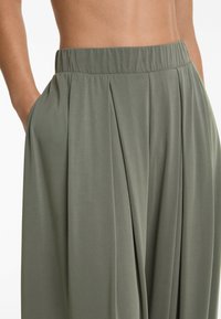 Pantaloni leggeri plissettati di color verde oliva con vita elastica e tasche laterali. Il tessuto ha una finitura liscia e leggermente strutturata.