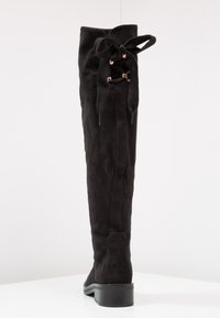 Botte haute en suède noir avec lacets à l'arrière et petit talon bloc sur une surface blanche avec un fond uni.