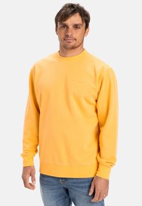 Sudadera amarilla de mangas largas y cuello redondo; presenta un logo texturizado en el pecho y puños y dobladillo acanalados. Llevada con jeans azules.