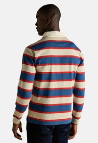 Mies, joka on käännetty pois, pukeutuneena pitkähihalaiseen rugby-paitaan, jossa on kerma, sininen ja punainen vaaka-raitakuosi, sekä tummiin housuihin.