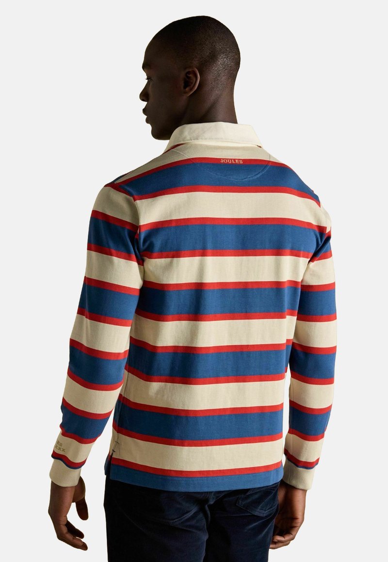 Mies, joka on käännetty pois, pukeutuneena pitkähihalaiseen rugby-paitaan, jossa on kerma, sininen ja punainen vaaka-raitakuosi, sekä tummiin housuihin.