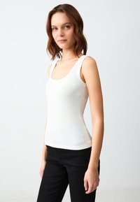 Giovane donna con capelli castani lunghi fino alle spalle che indossa una maglietta bianca senza maniche e pantaloni neri su uno sfondo chiaro e uniforme.