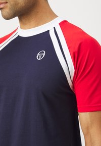 T-shirt in cotone navy con maniche corte rosse, bordature bianche e un logo circolare sul petto sinistro; presenta un design sportivo con colori a contrasto.