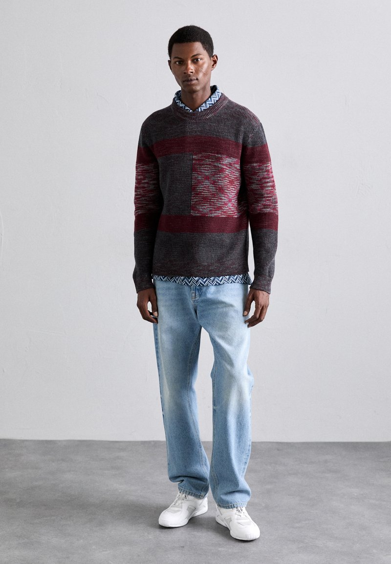 Dark gray and burgundy patterned sweater layered over a blue patterned shirt, paired with light blue jeans and white sneakers.