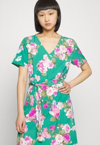 Vestido floral com decote em V, mangas curtas e um cinto na cintura. Feito de tecido verde com grandes flores rosa, brancas e roxas.