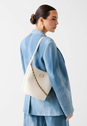 Frau mit hochgesteckten Haaren, die goldene Creolen und einen blauen Anzug trägt, eine weiße Handtasche mit goldener Kette auf der Schulter vor weißem Hintergrund.