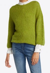 Groene gebreide trui met een rechthoekige vorm, brede mouwen en een gestructureerde afwerking. Draagt over een witte blouse met ruches, gecombineerd met een blauwe spijkerbroek.