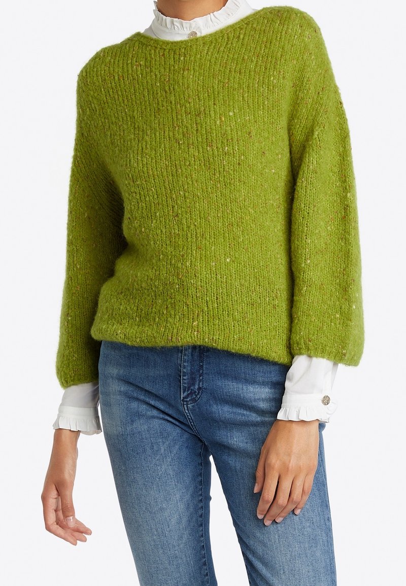 Groene gebreide trui met een rechthoekige vorm, brede mouwen en een gestructureerde afwerking. Draagt over een witte blouse met ruches, gecombineerd met een blauwe spijkerbroek.