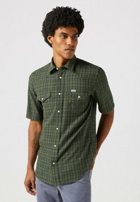Chemise à manches courtes à carreaux verts avec des boutons blancs et deux poches poitrine. Étiquette Wrangler sur la poche gauche. Tissu doux et texturé.