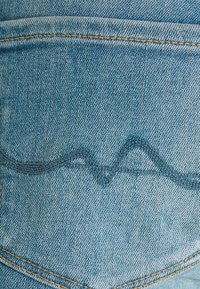 Vaaleansininen farkkukangas, jossa on tekstuuri, ja tummansinisellä langalla ommeltu aaltoileva kuvio sekä erottuva keltainen sauma.