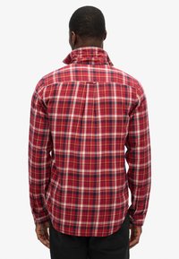 Camicia a quadri rossa con accenti bianchi e blu navy, maniche lunghe, polsini con bottoni, colletto alto e orlo dritto. Dettaglio importante della piega posteriore.