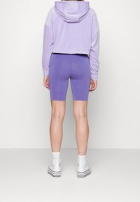 Cropped hoodie viola con cappuccio, abbinata a pantaloni da ciclista viola coordinati. Il modello indossa sneaker bianche alte, mostrando texture morbide.