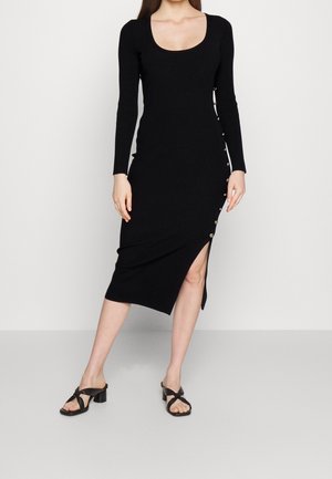 Femme portant une robe midi noire à manches longues avec des boutons sur le côté et une fente haute sur la cuisse, associée à des sandales noires à talons.