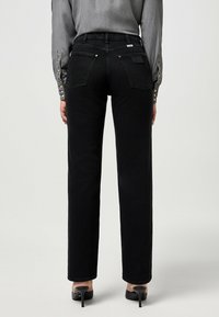 Vaqueros negros de cintura alta con pierna recta, costuras en contraste, dos bolsillos traseros y una etiqueta en la cinturilla, combinados con una camiseta gris.