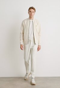 Cream zip-up bomber jacket with a subtle logo, paired with matching joggers. Footwear is light gray. Minimalist design and fit.