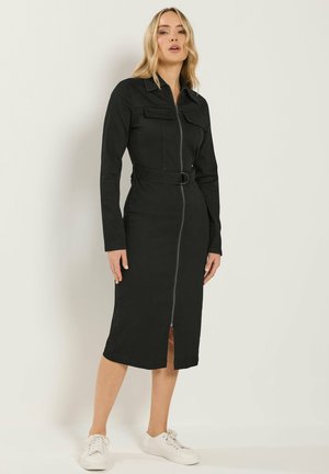 Kobieta stojąca, ubrana w czarną, midi sukienkę z długim rękawem, z suwakiem z przodu i paskiem, w połączeniu z białymi trampkami na jednolitym jasnym tle.