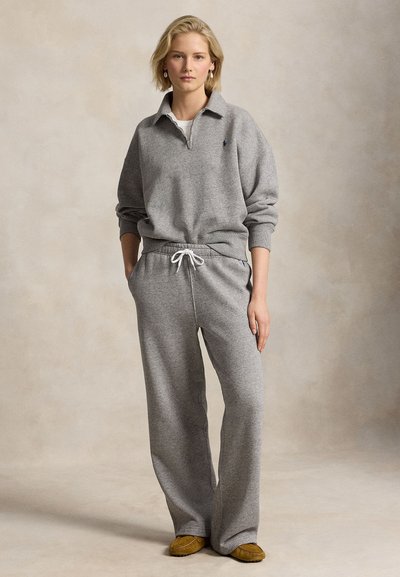 Blonde vrouw staat, draagt een grijze kwart-ritssluiting sweater en bijpassende joggingbroek met witte trekkoord en beige mocassins.