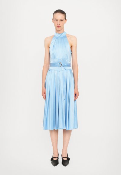 Abito halter in satin azzurro chiaro con gonna plissettata e vita cinturata con una fibbia circolare. Il modello indossa scarpe nere con punta a forma di stiletto.