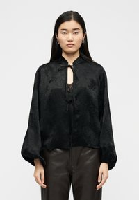 Νεαρή γυναίκα με μακριά σκούρα μαλλιά φοράει μια μαύρη υφασμάτινη μπλούζα με δέστρες και δερμάτινο παντελόνι, στέκεται μπροστά σε απλό φόντο.