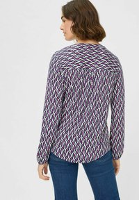 Langärmlige Bluse in Weiß mit einem lila und rosa wellenförmigen Muster. Verfügt über einen runden Ausschnitt und einen gesammelten Rücken für zusätzliche Textur.